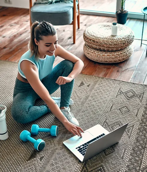 A woman in sportswear sitting on a rug next to a pair of dumbbells, a water bottle, and furniture, while looking at her laptop.