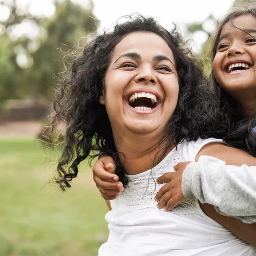 A mother and her young daughter laughing in a green field while the daughter holds onto her mother’s shoulders.