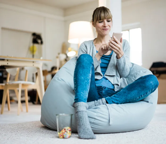 A woman in blue jeans looking at her smartphone while sitting casually on a bean bag chair in the middle of a bright, modern room.