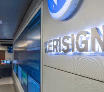 A closeup of a Verisign logo sign which is lit-up on an office wall that’s next to a big digital screen showing multiple dashboards.