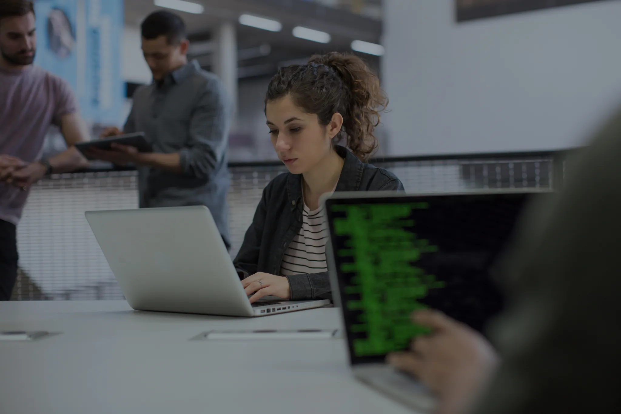 Professionals in an office standing and talking over a tablet while a woman sitting at a laptop types up a report.