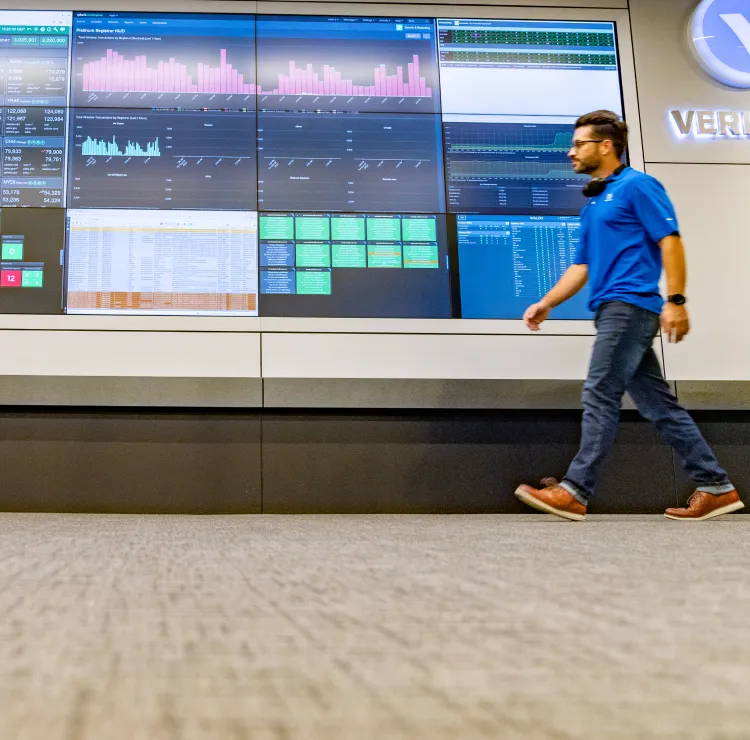 A man walking in front of a big display of screens showing bar graphs and detailed reports.