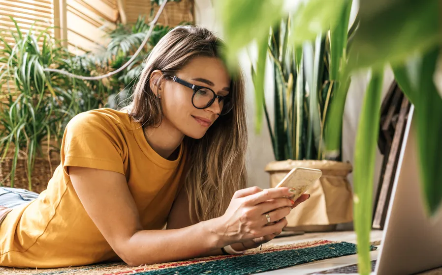 A young woman relaxing at a home with many plants around her as she’s lying on the floor and working on her smartphone.