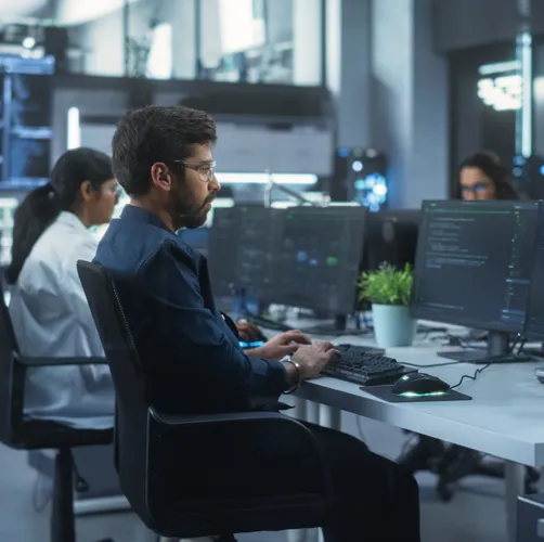 Professionals working on technological research and development projects on desktop computers at a shared table in an office.