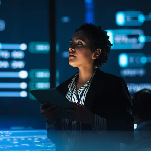 A professional woman uses a tablet computer in a darkened system control monitoring center with multiple lighted displays.