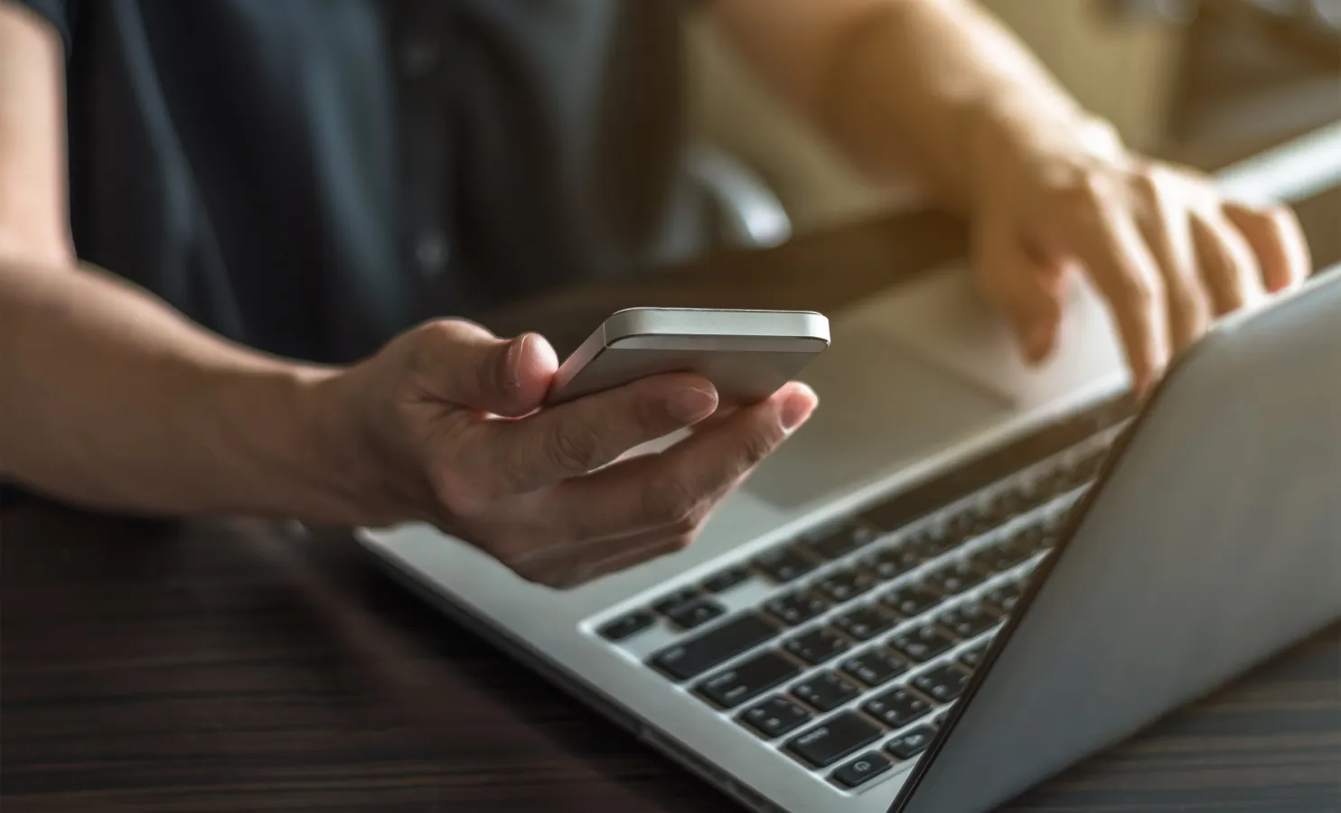 Closeup of a professional holding a smartphone in one hand and typing on a laptop with the other hand.