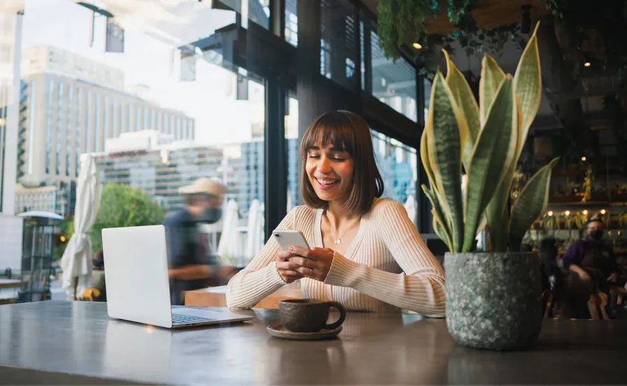 A woman looking at her smartphone and sitting in a casual café in front of her laptop, a coffee mug, and next to a plant.