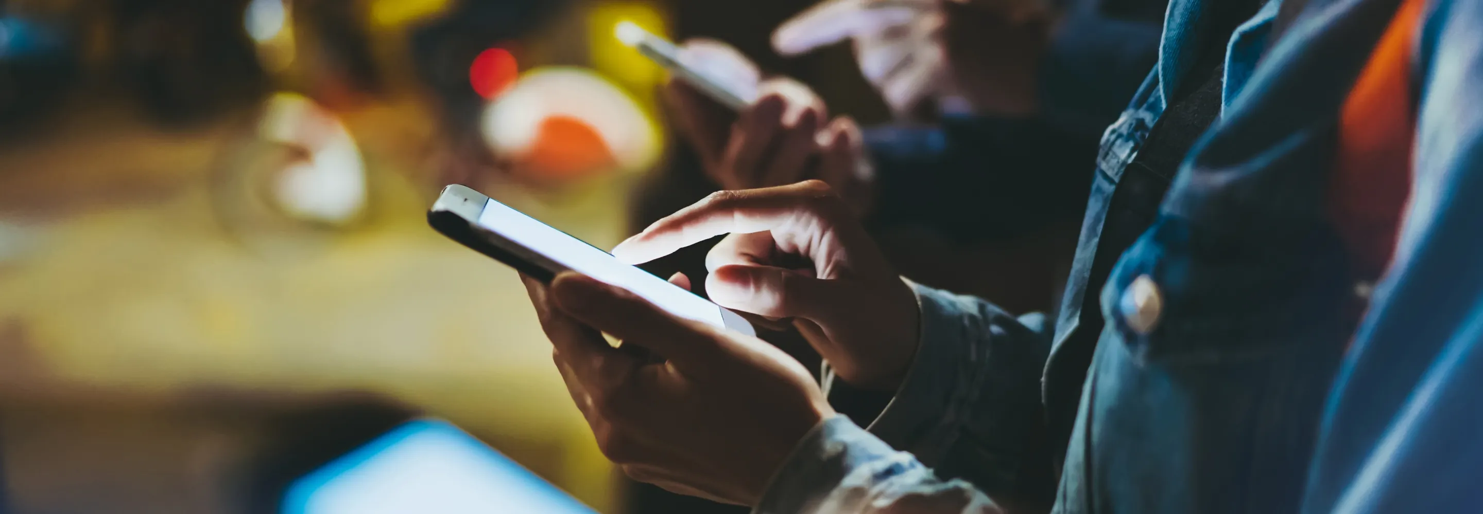 Close shot of people’s hands scrolling through their smartphones that light up at night with city lights in the background.