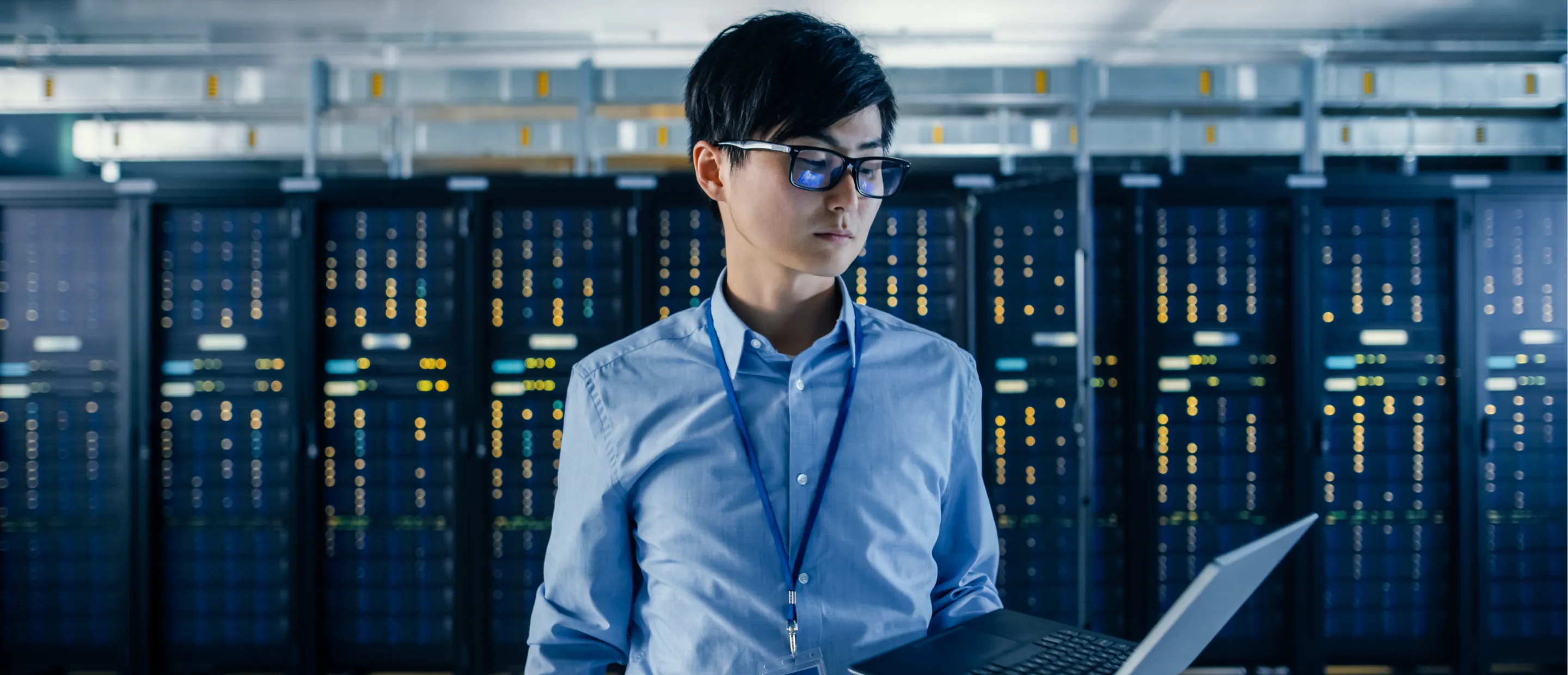 A professional man wearing an ID badge standing in front of a large data server while he works on a laptop.