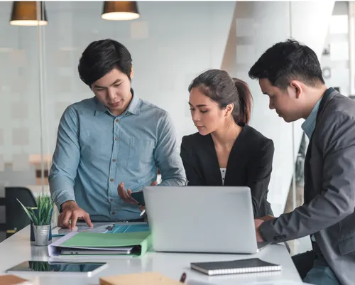 Three professionals in an office setting work together with a laptop and binders.