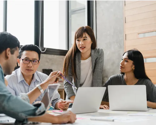 Four colleagues brainstorm together with their laptops in an office meeting room that has a big window with natural light.